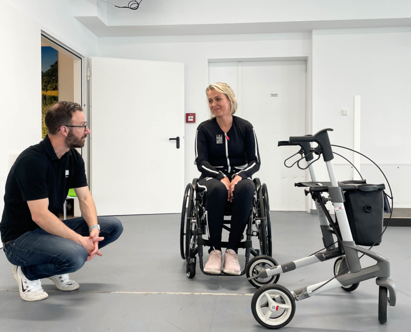 Patientin sitzt im Rollstuhl, ein rahm Mitarbeiter kniet links von ihr